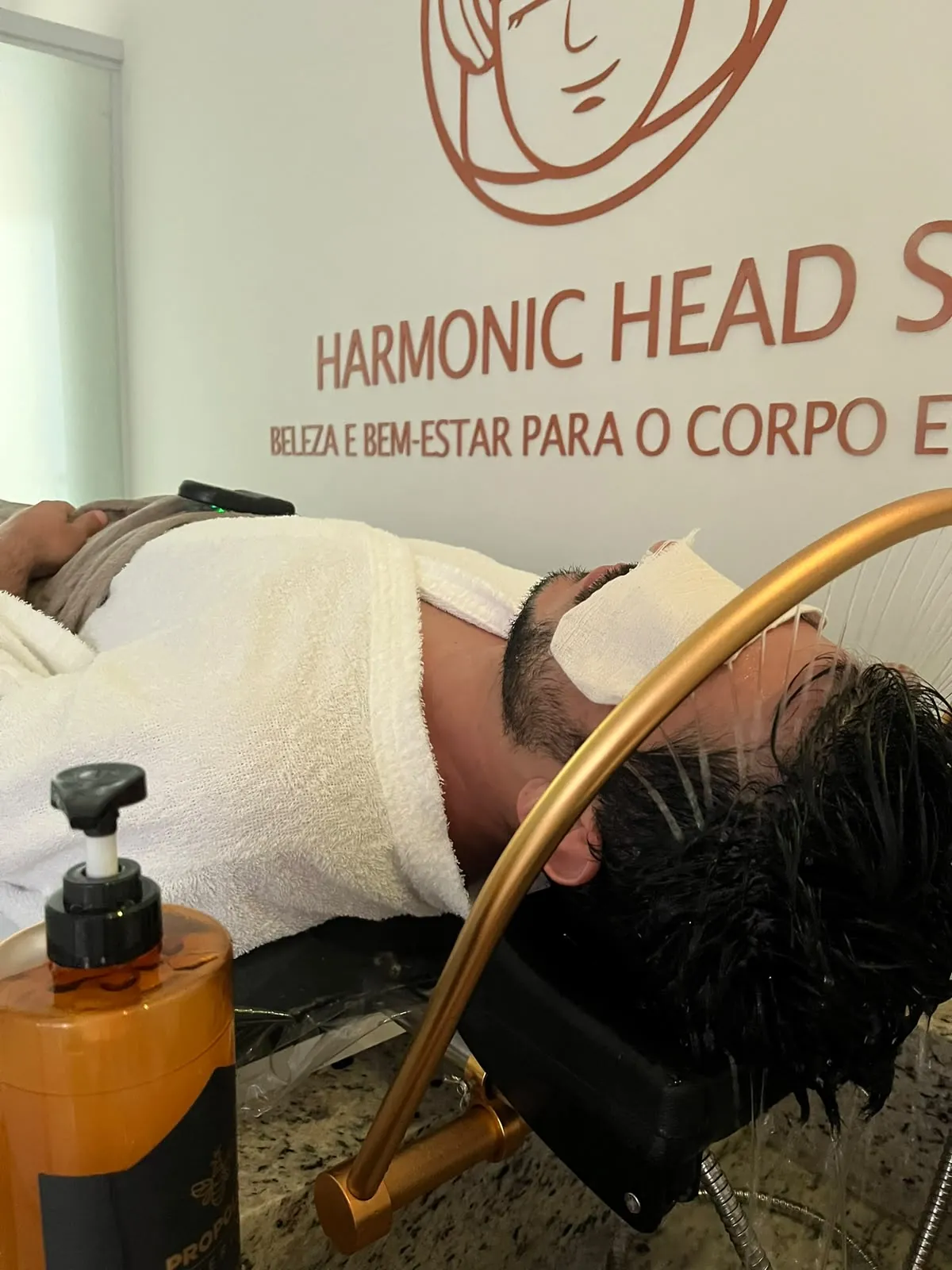 Cliente com máscara nos olhos sob o arco dourado, com a logo Harmonic Head Spa na parede